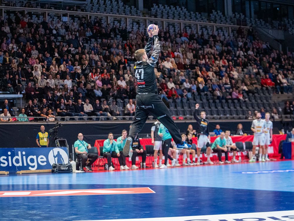 Handball players compete in an intense indoor match with spectators watching from the stadium stands.