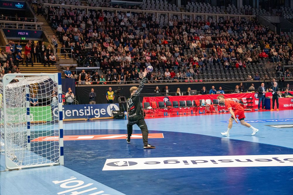 Indoor handball match showing a penalty shot attempt with players positioned around the court.