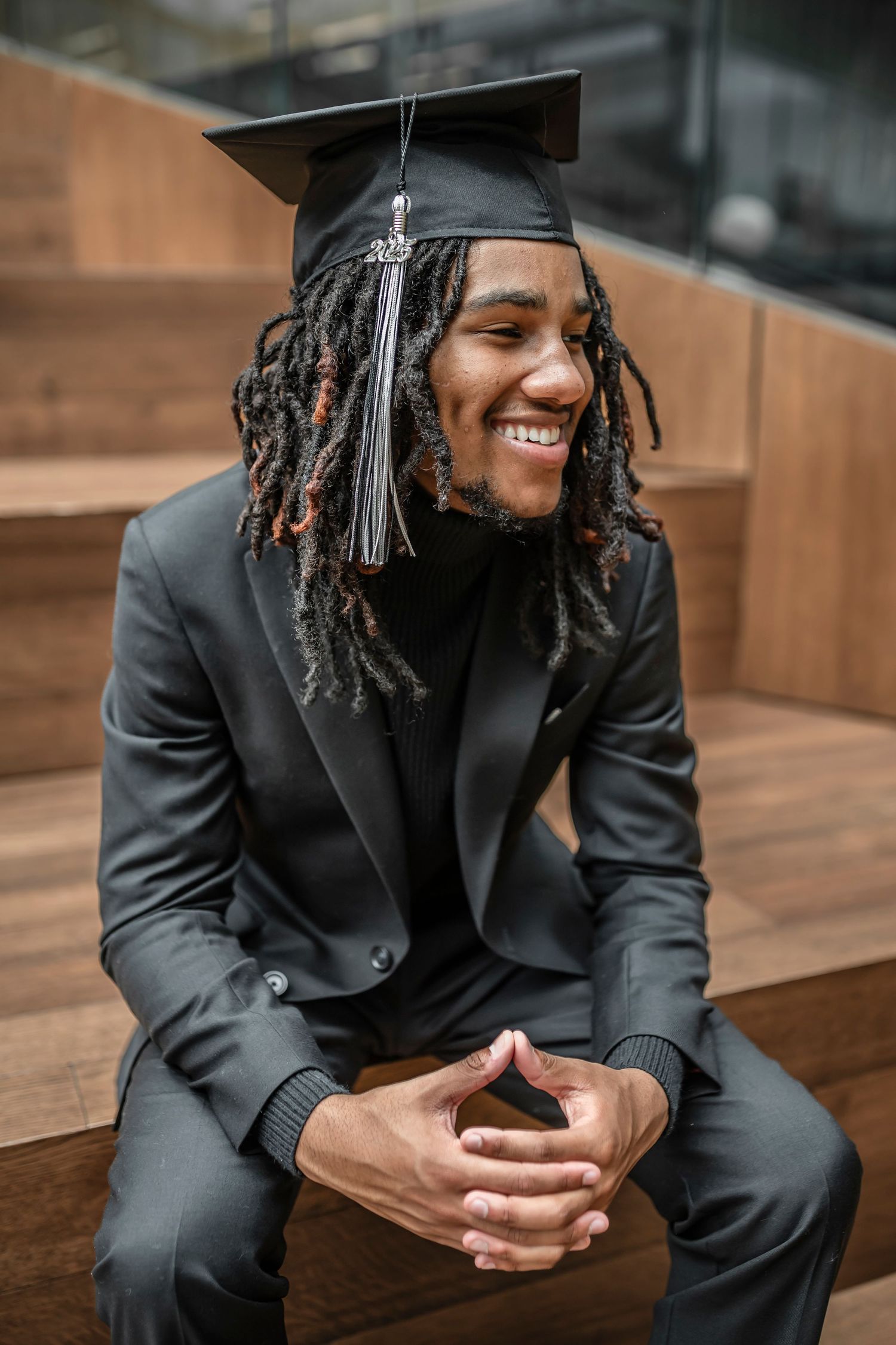 Graduate wearing black academic regalia with purple hood stands against brick wall in outdoor portrait.