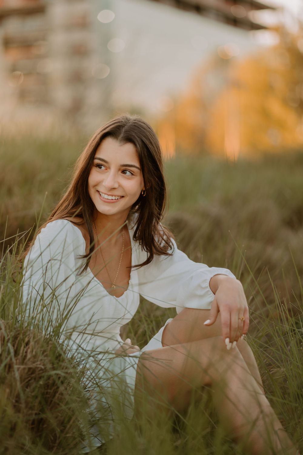 Smiling woman in a white dress sits in a grassy field during golden hour.