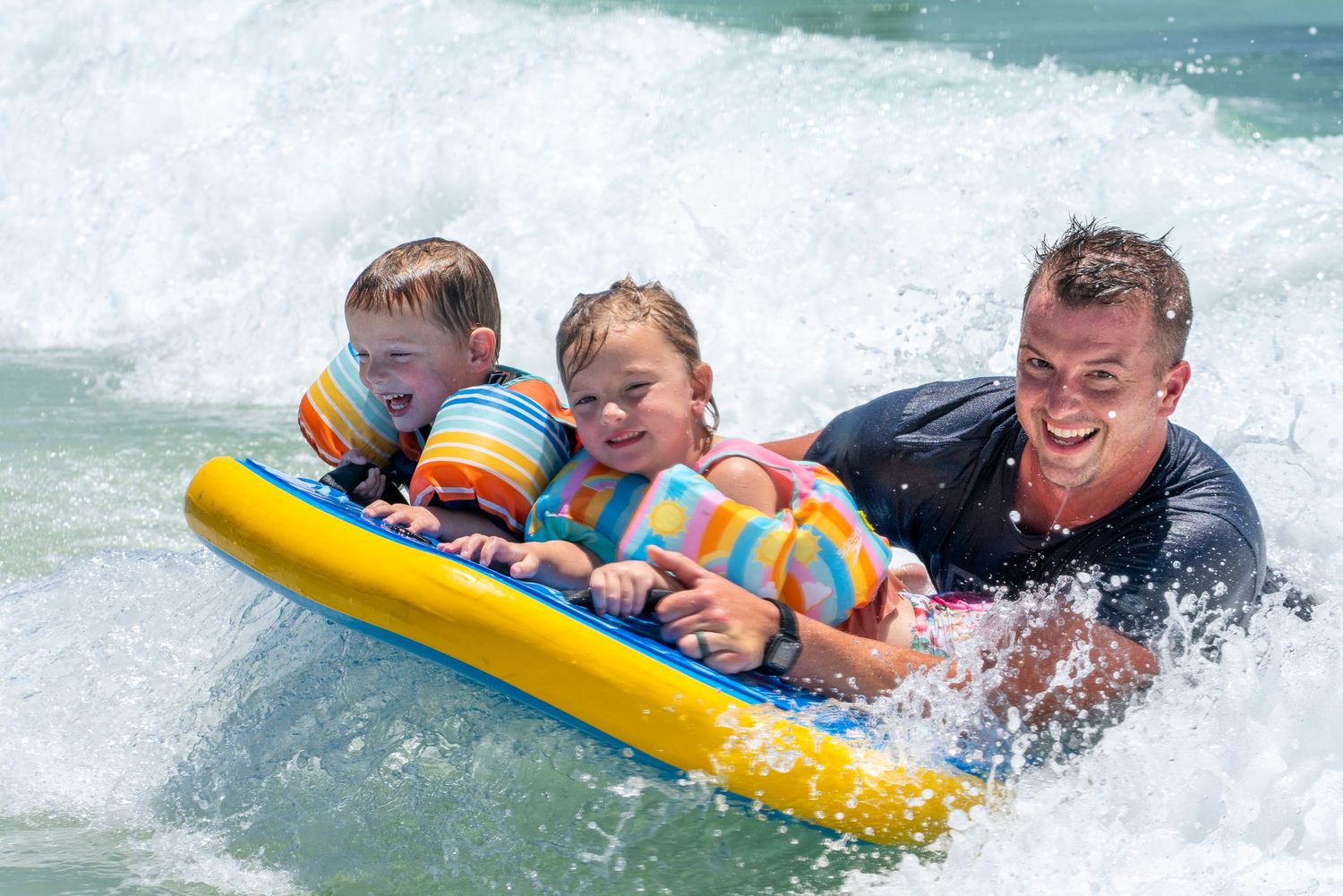 Dad and children riding waves on colorful boogie boards in white ocean surf.