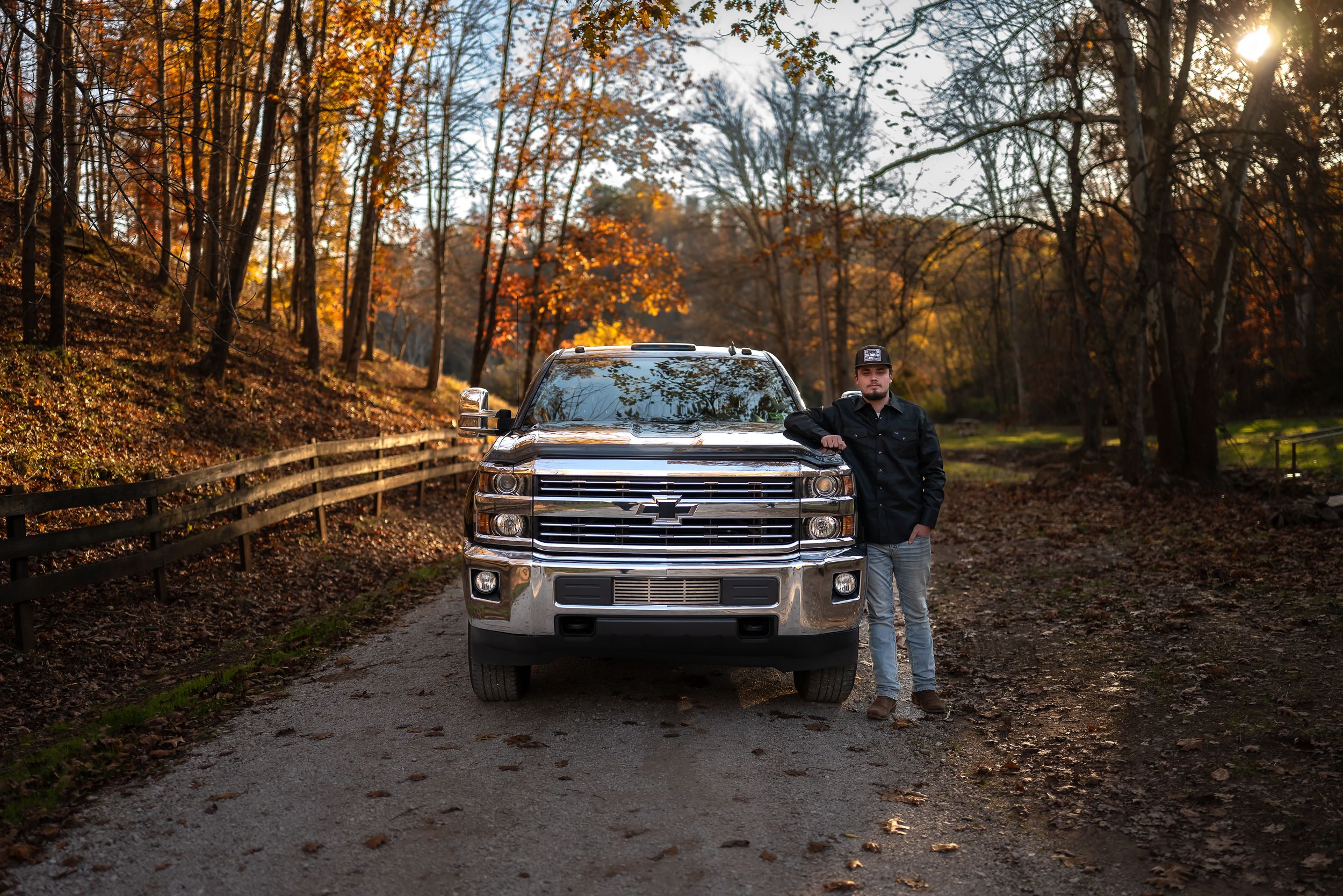 A vintage car is parked on a forest trail during autumn with golden sunlight filtering through the trees.