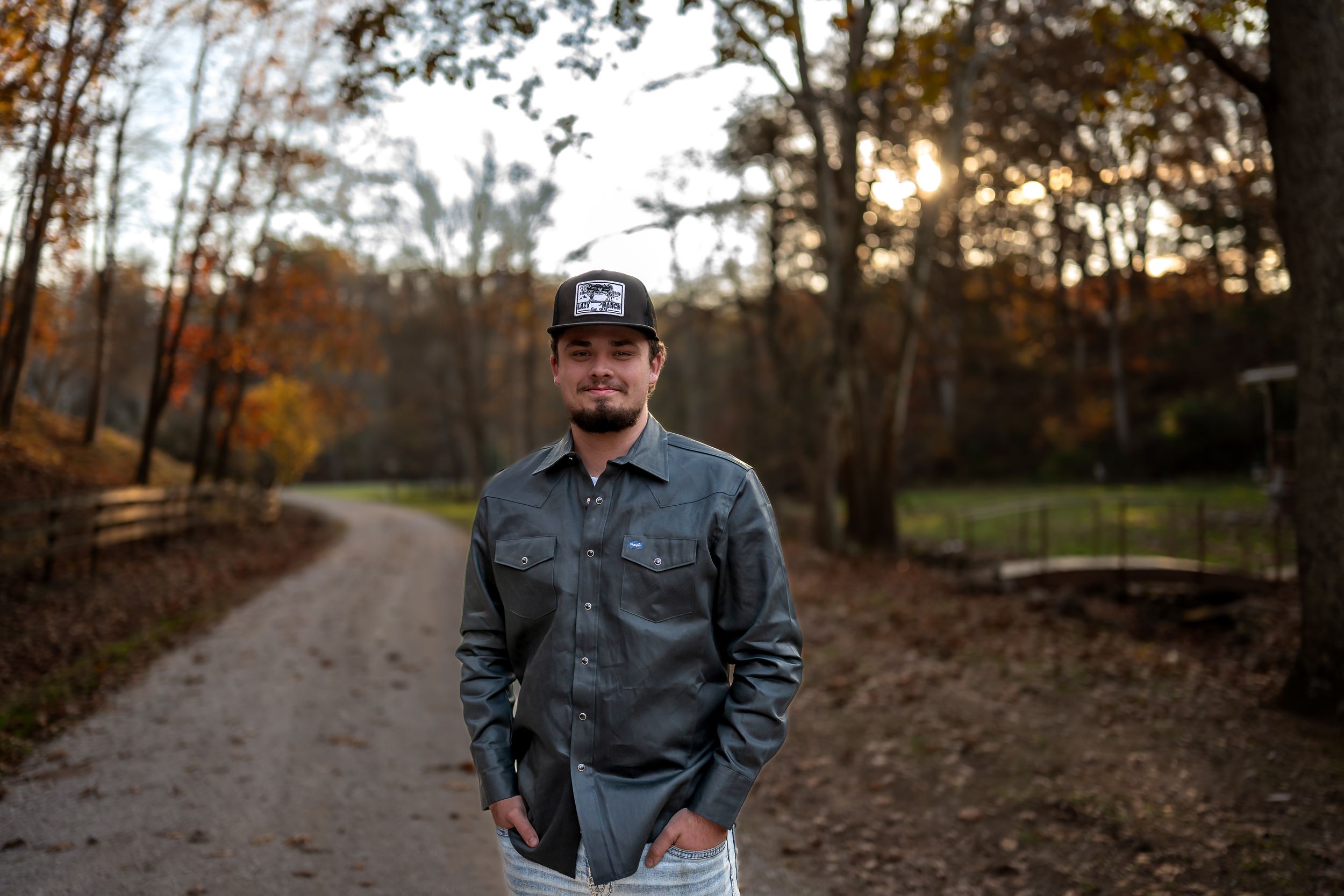 Person in denim jacket poses against autumn forest backdrop during golden hour sunset.