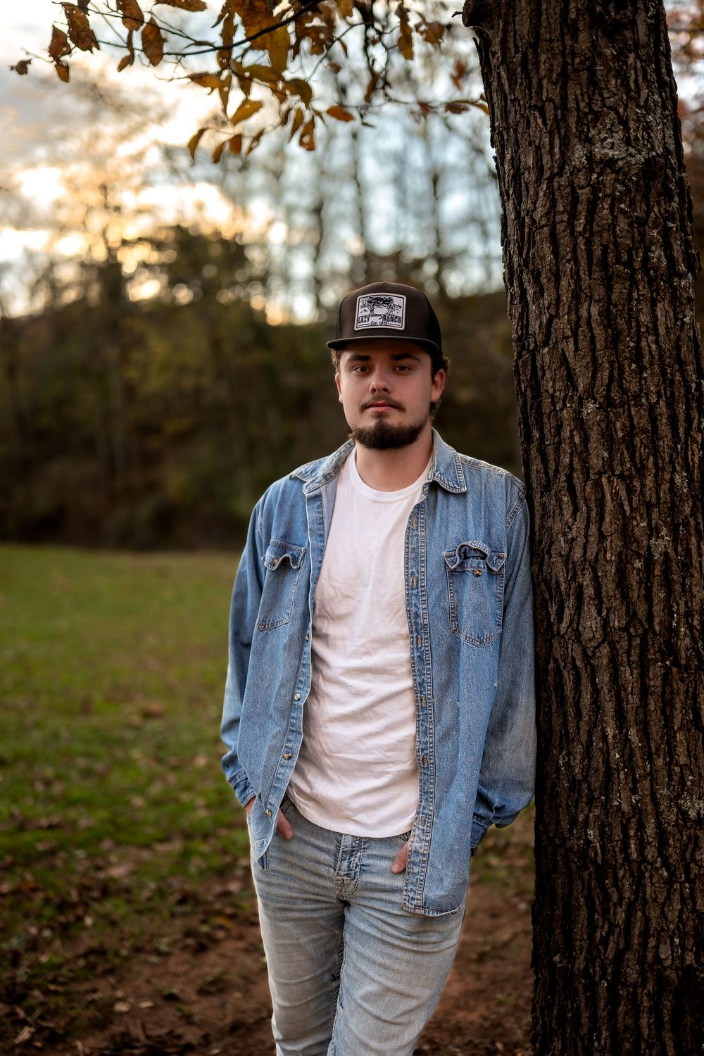 Person in denim jacket and baseball cap stands against tree trunk in autumn forest setting.