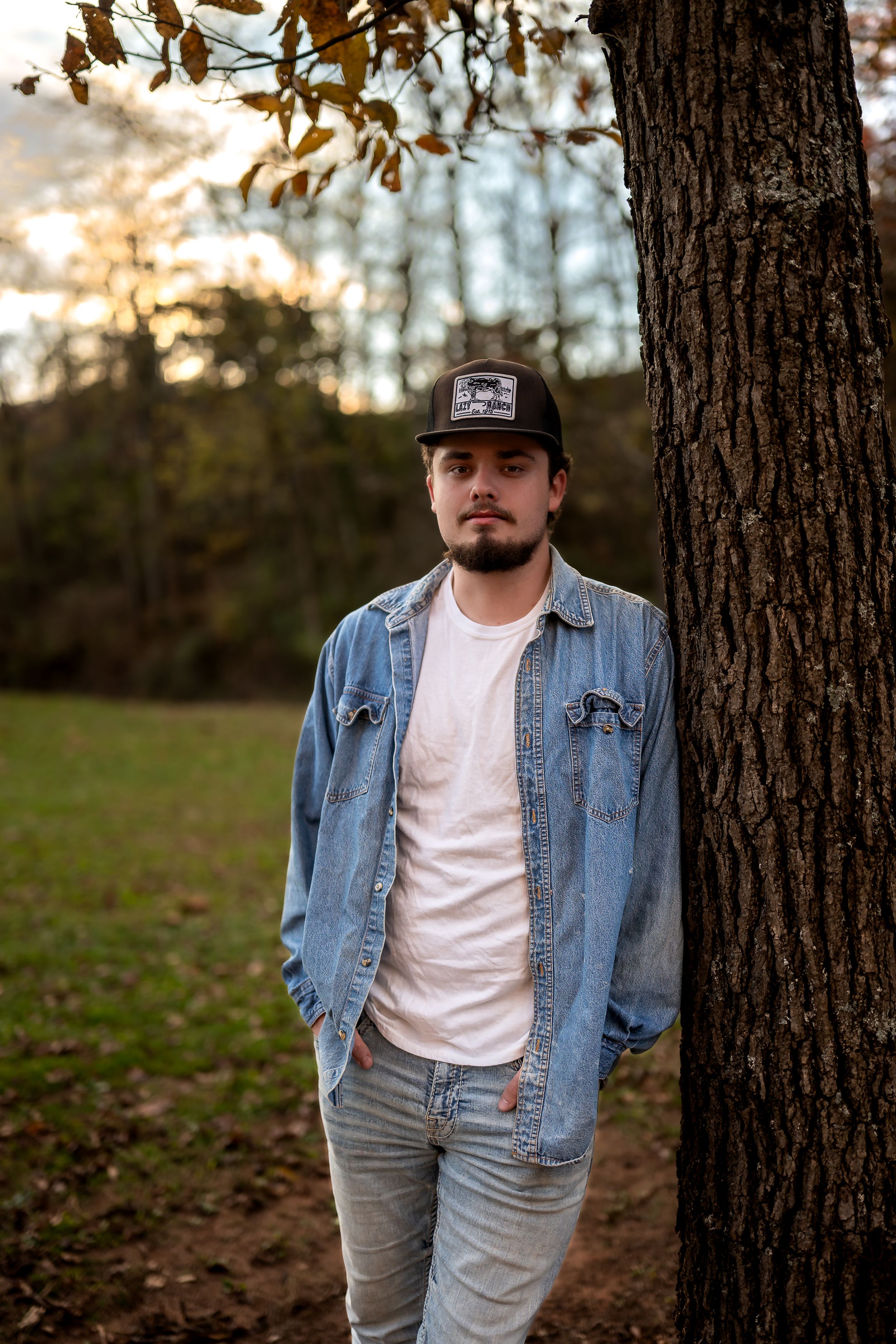 Person in denim jacket and baseball cap stands against tree trunk in autumn forest setting.