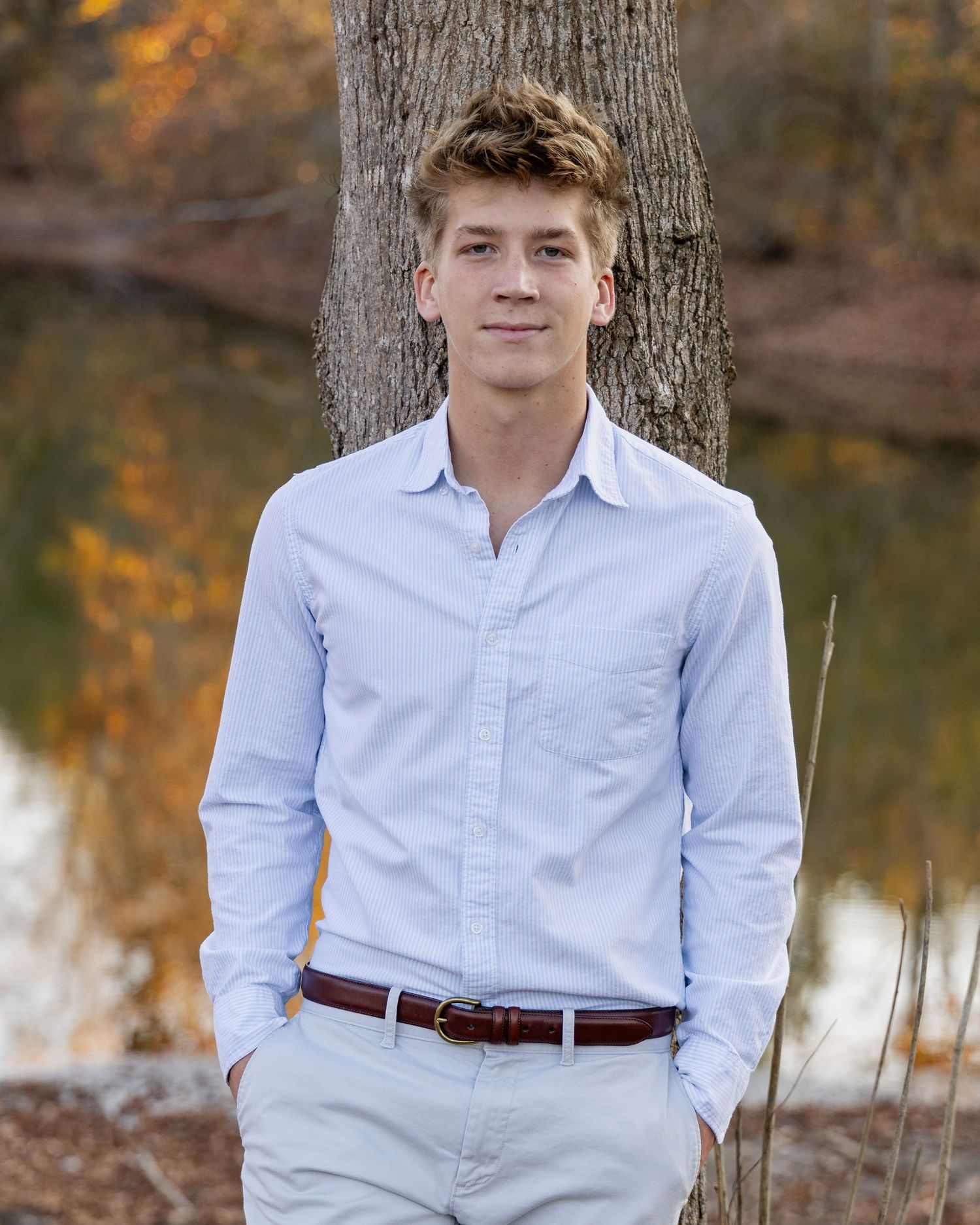 Person in light blue button-down shirt sitting against tree trunk near water.