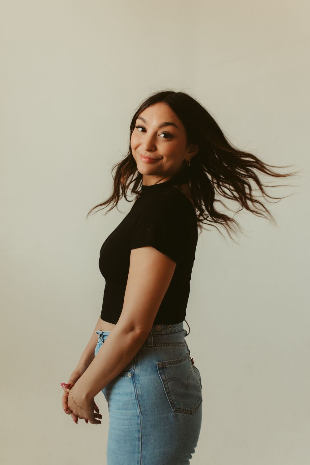 Young woman in black crop top and jeans posing with hair flowing, smiling over shoulder against light background.