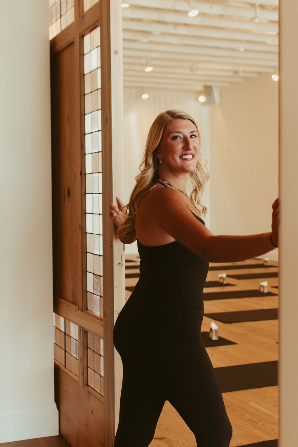Smiling blonde woman in black athletic wear poses in doorway of modern yoga studio with wood floors.