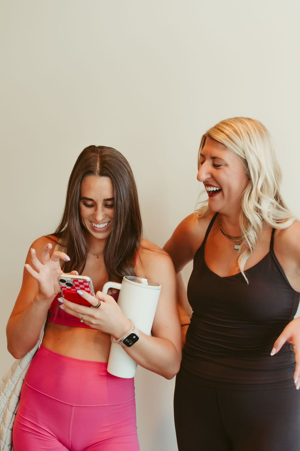 Two women in athletic wear laugh together while looking at a smartphone, one in pink and one in black activewear.