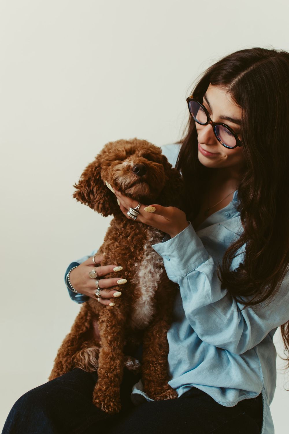 Young woman with glasses holding a fluffy brown miniature poodle puppy against a light background.