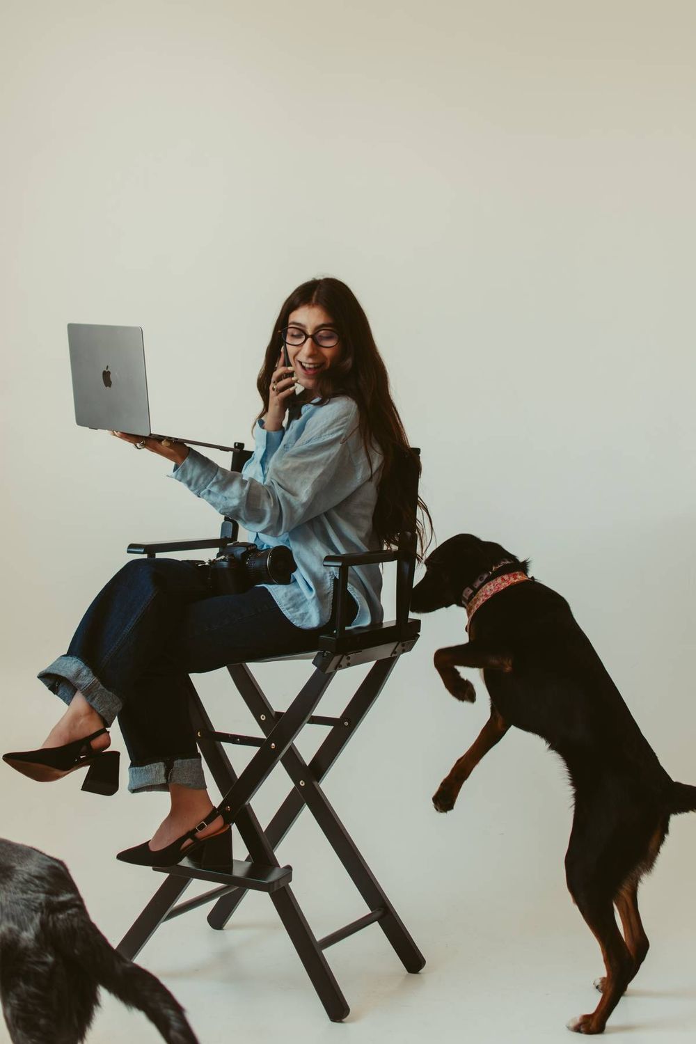 Woman sitting in director's chair holding Apple laptop and phone while black dog jumps up beside her in studio setting.