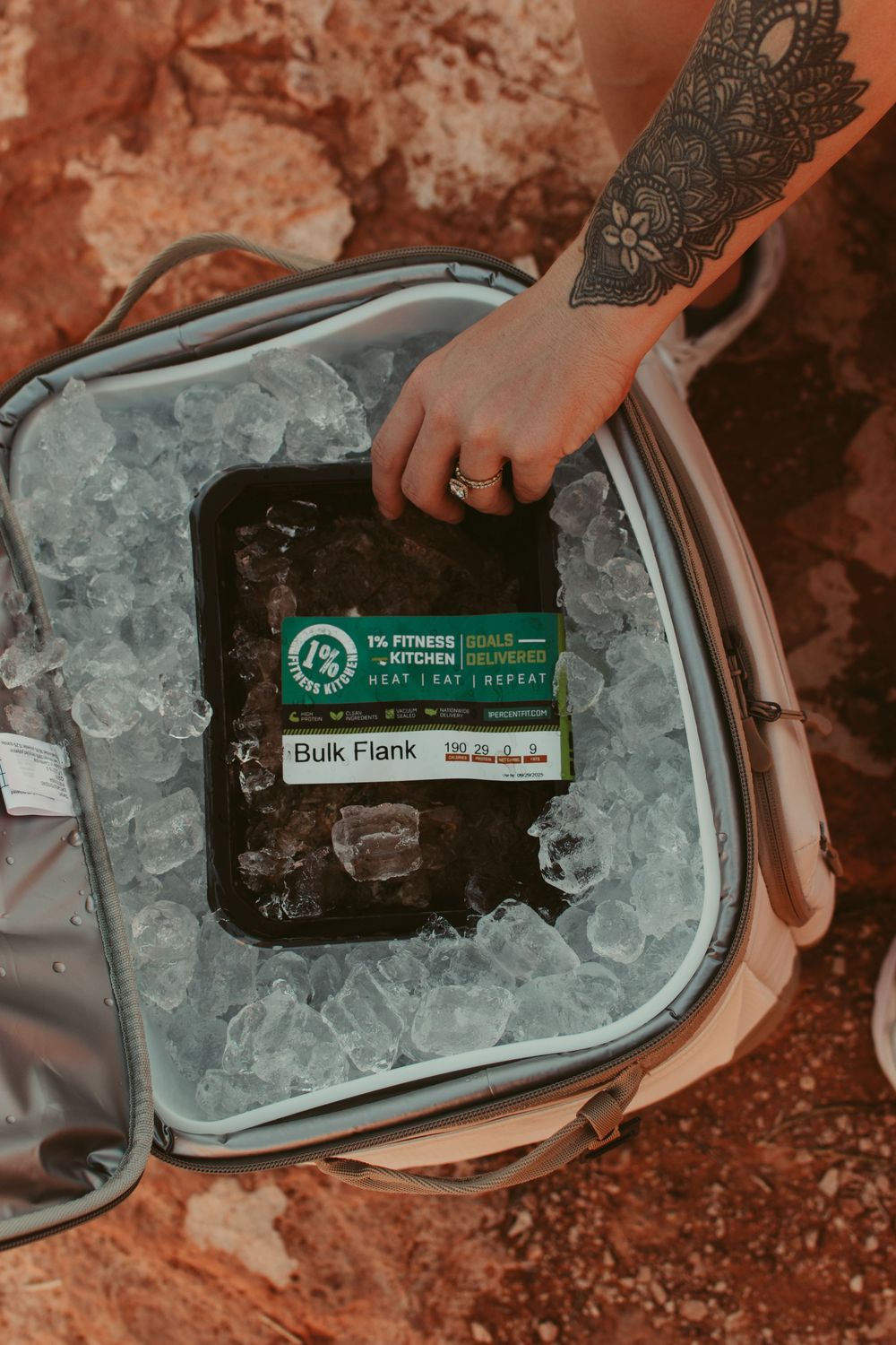 Tattooed hand placing bulk flank meat into an ice-filled cooler bag outdoors on rocky terrain.