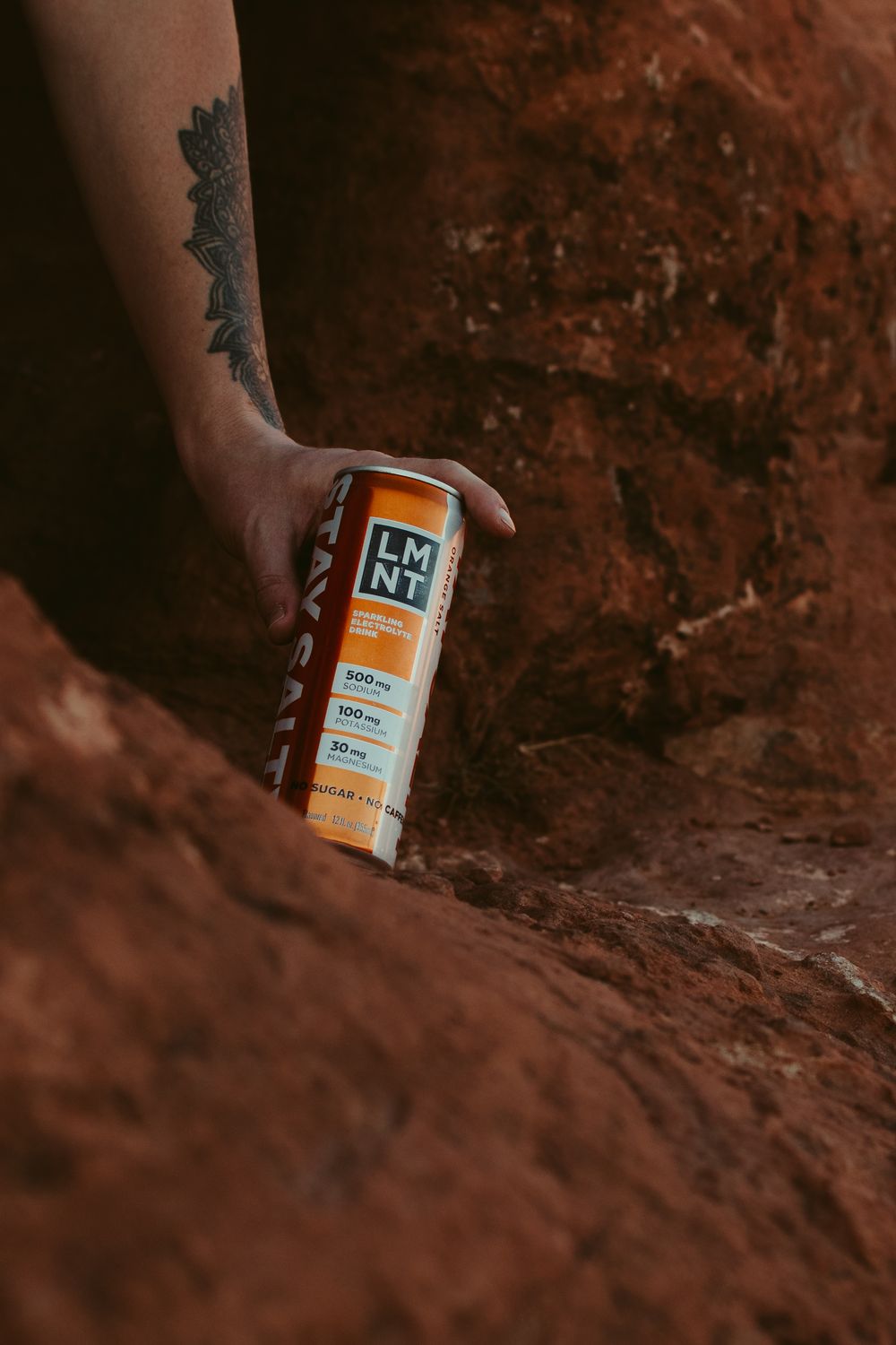 Tattooed hand holding LMNT electrolyte drink can against red rock canyon wall outdoors.