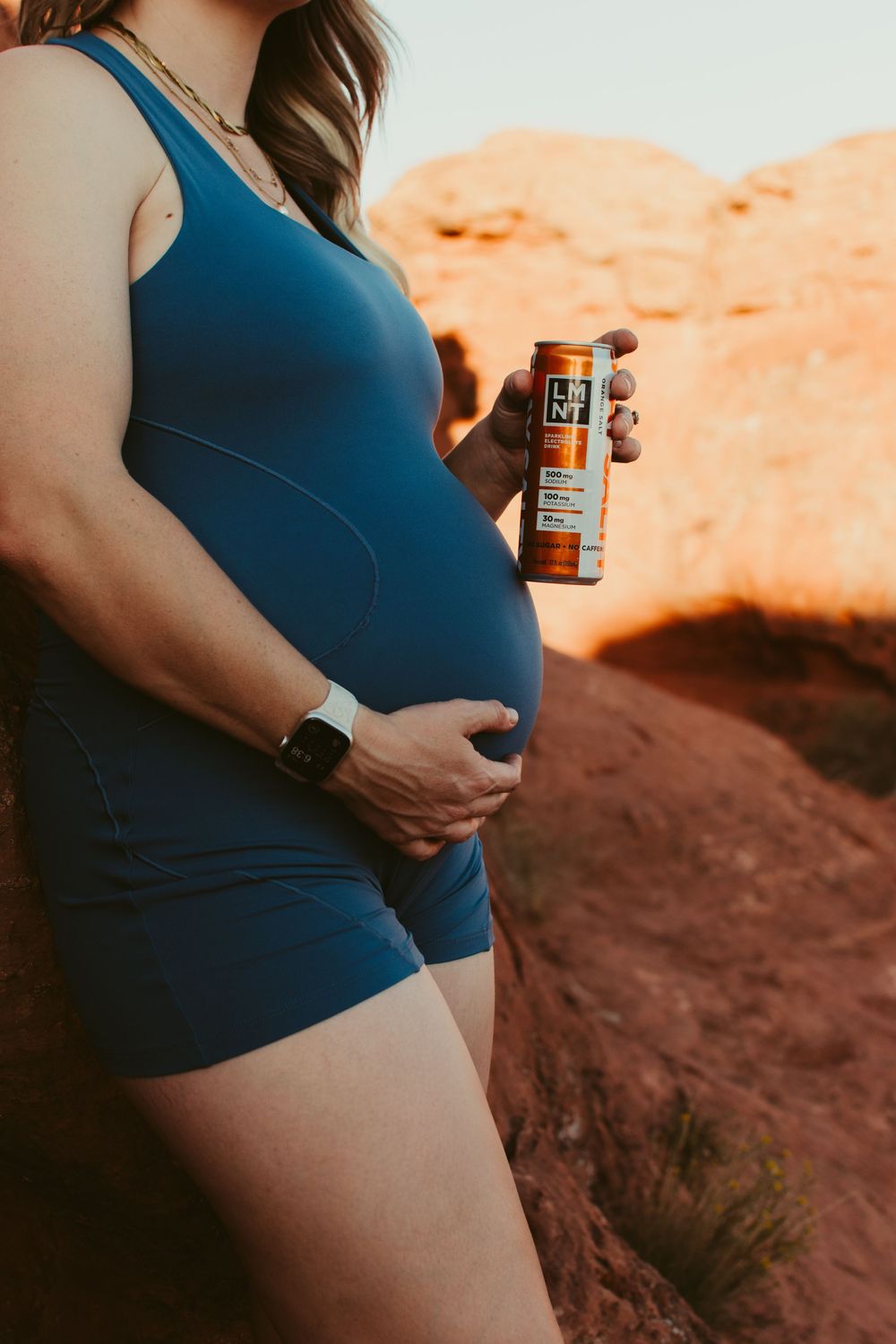 Pregnant woman in blue romper holding an energy drink can outdoors near red rock formations.