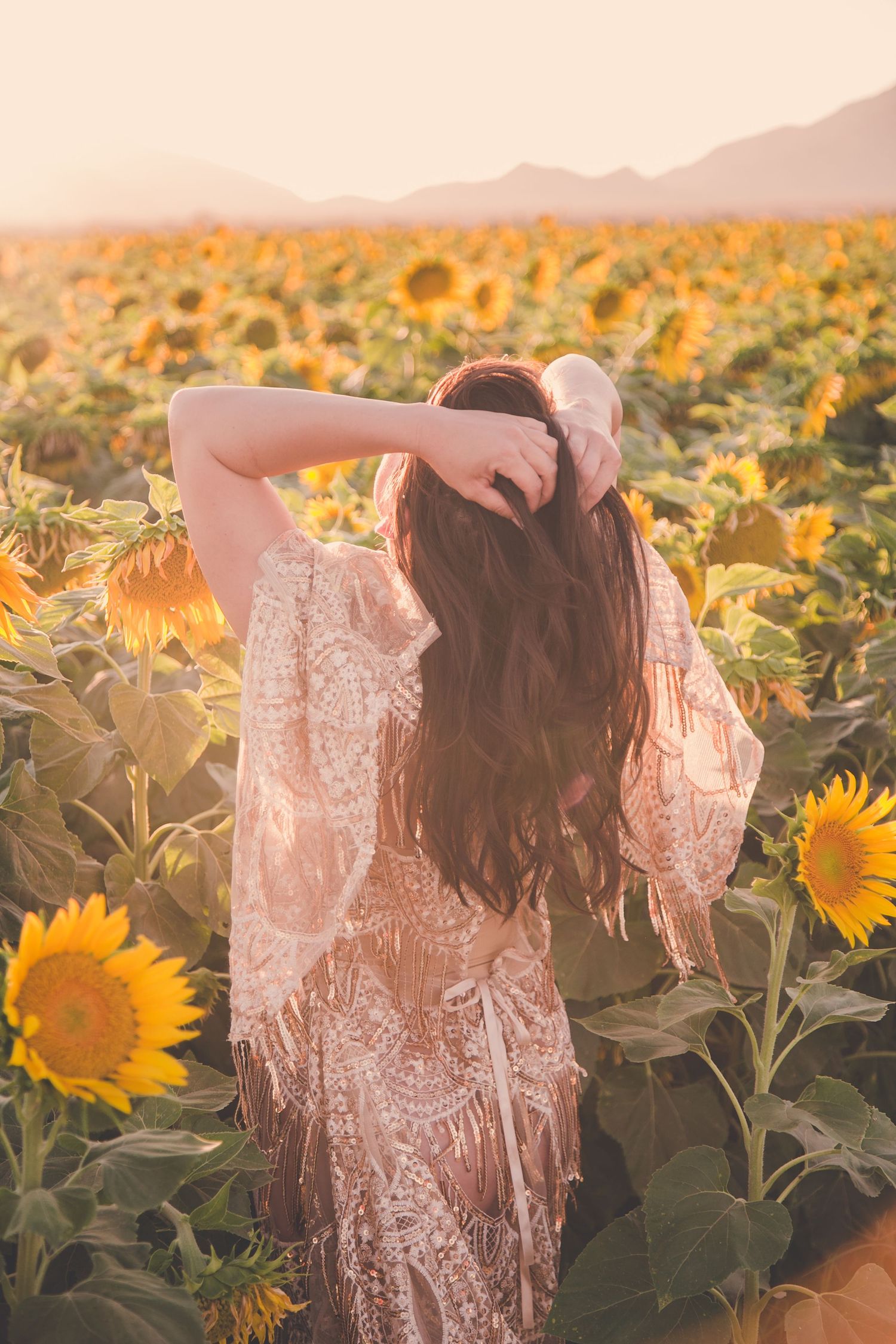 Woman in lace dress standing in sunflower field at golden hour with mountains in background.