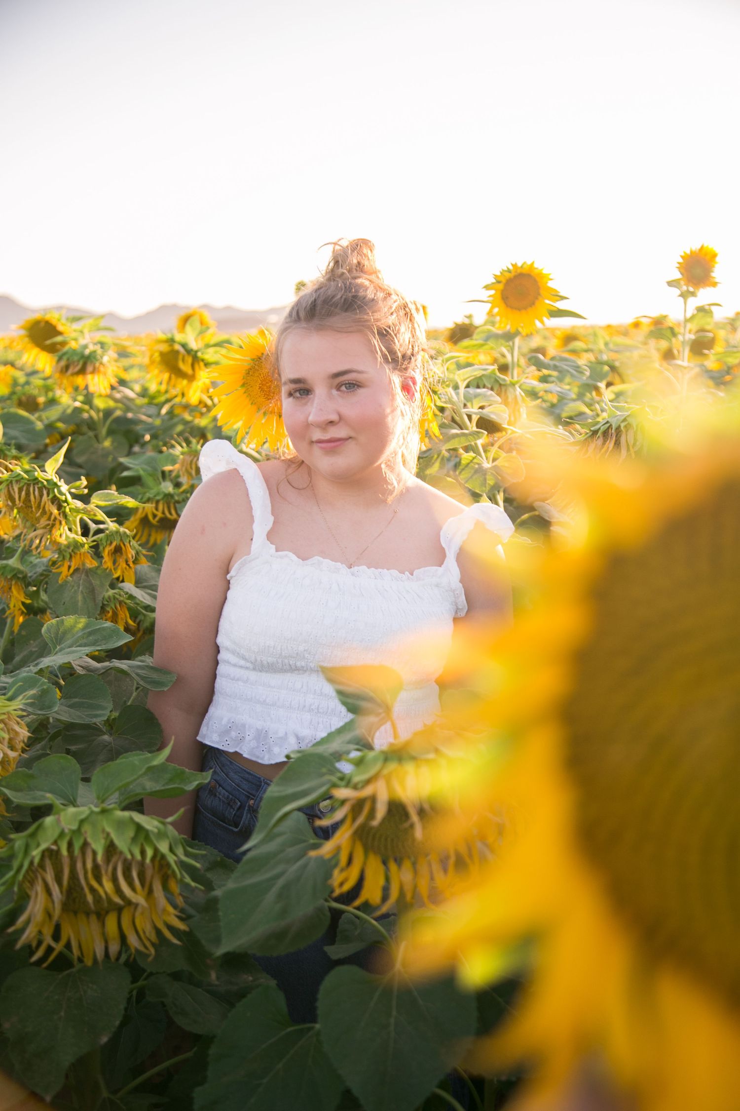 Young woman in white crop top surrounded by tall sunflowers in a glowing sunlit field.