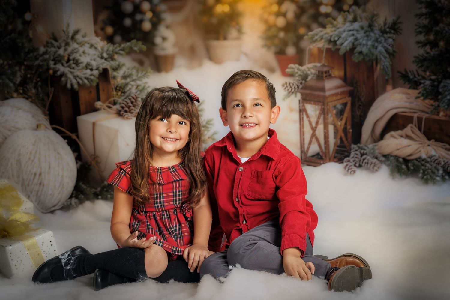 Two young children in red holiday outfits pose together in a festive Christmas photo studio setting.