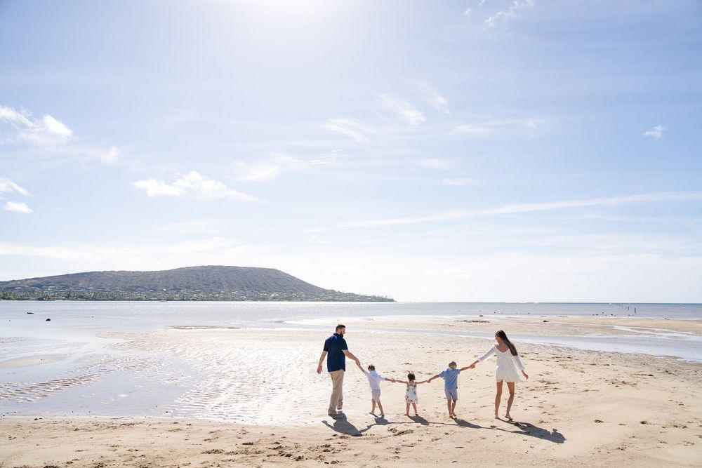 People walking hand in hand along a sandy beach with ocean waves and mountains in the background.