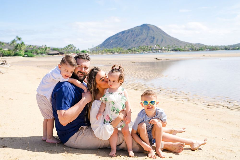 A family sits together on a sandy tropical beach during a sunny day with palm trees and mountains in the background.