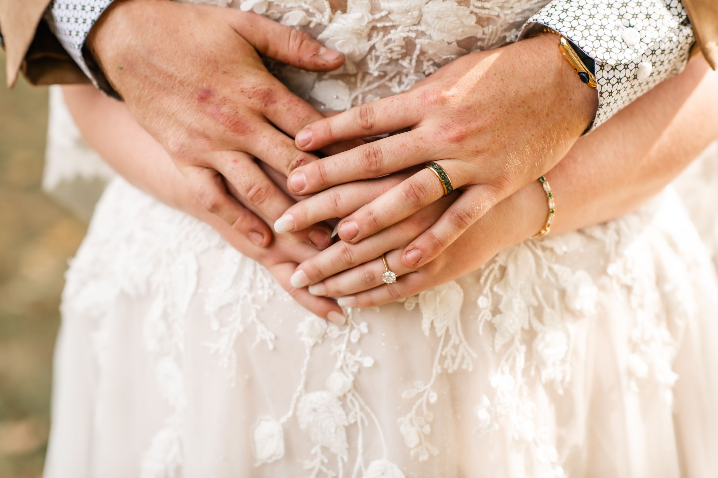 Close-up of hands with wedding rings resting on white lace dress.
