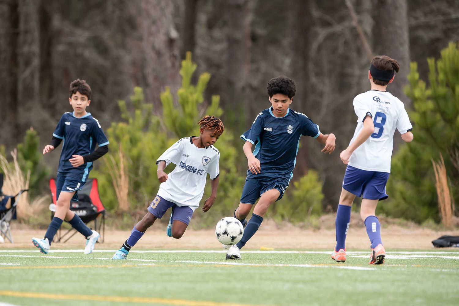 Youth soccer players in white and blue uniforms battling for the ball on a grass field.