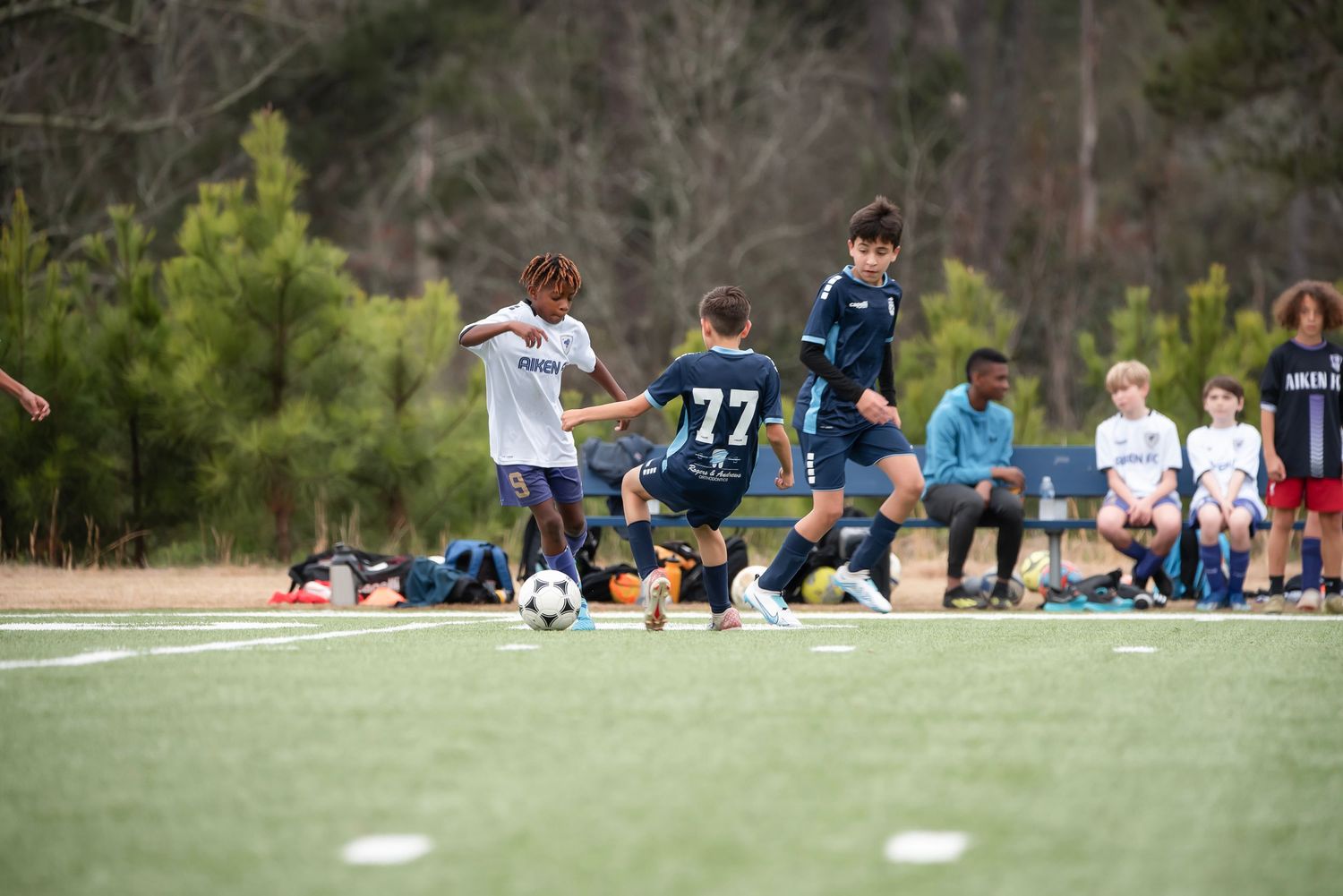 Soccer teams in blue and white uniforms playing on a field with spectators watching from sidelines.
