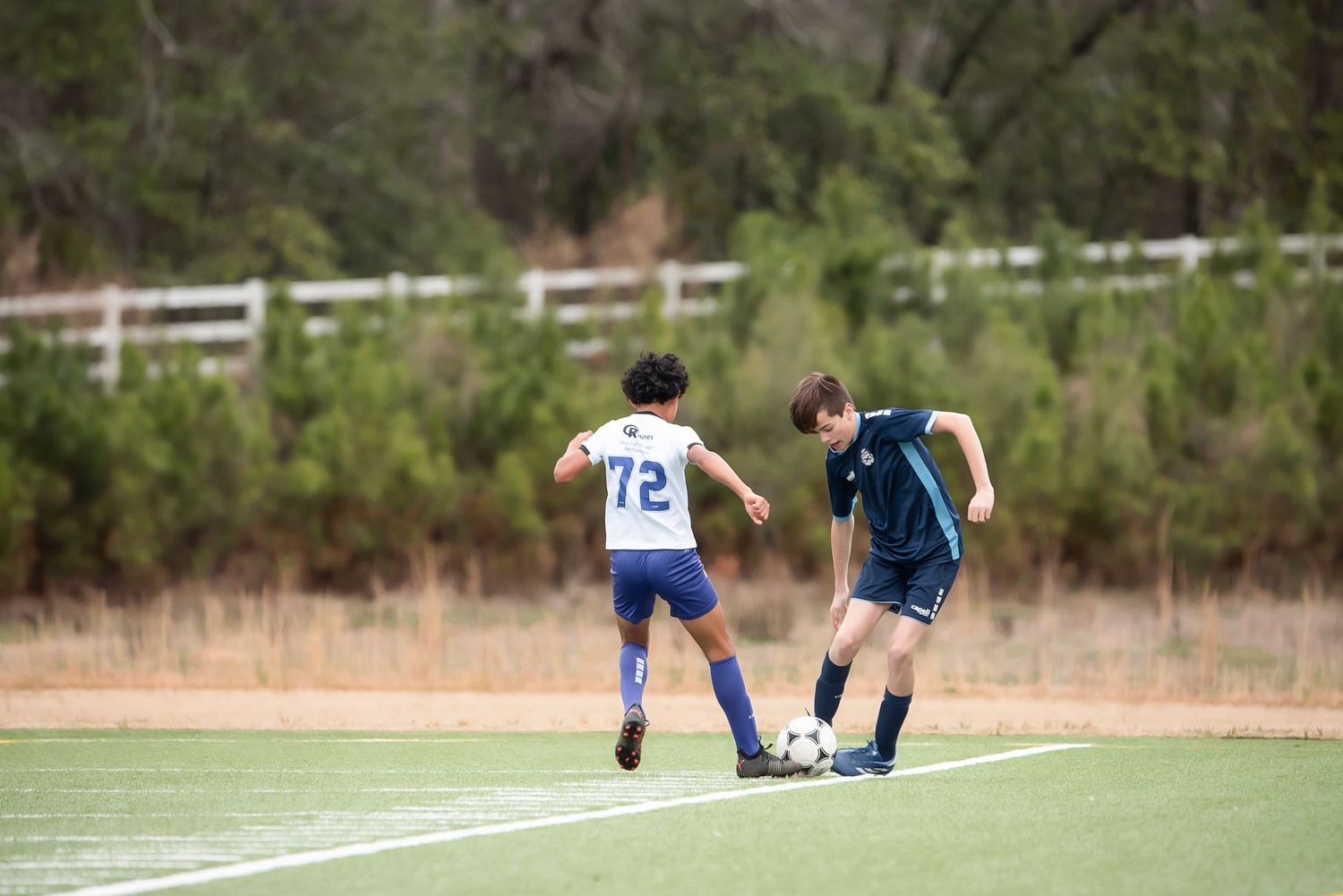 Soccer players spread out across a turf field during a match with trees in the background.