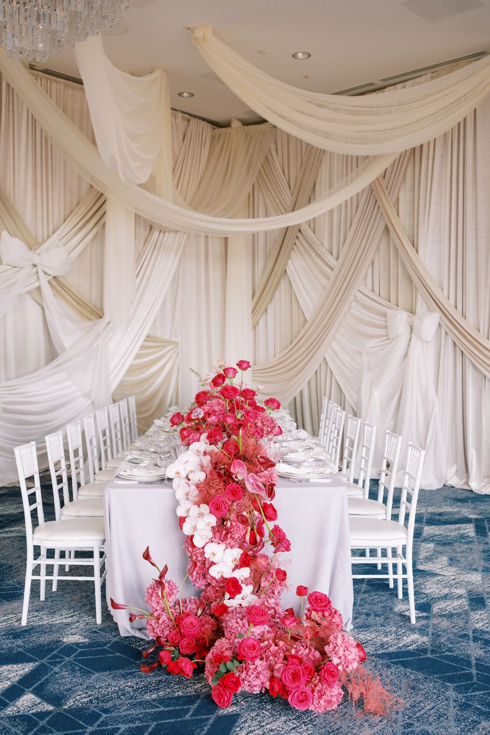 Elegant wedding table setup with dramatic white drapes and cascading pink floral arrangement on blue carpeted floor.