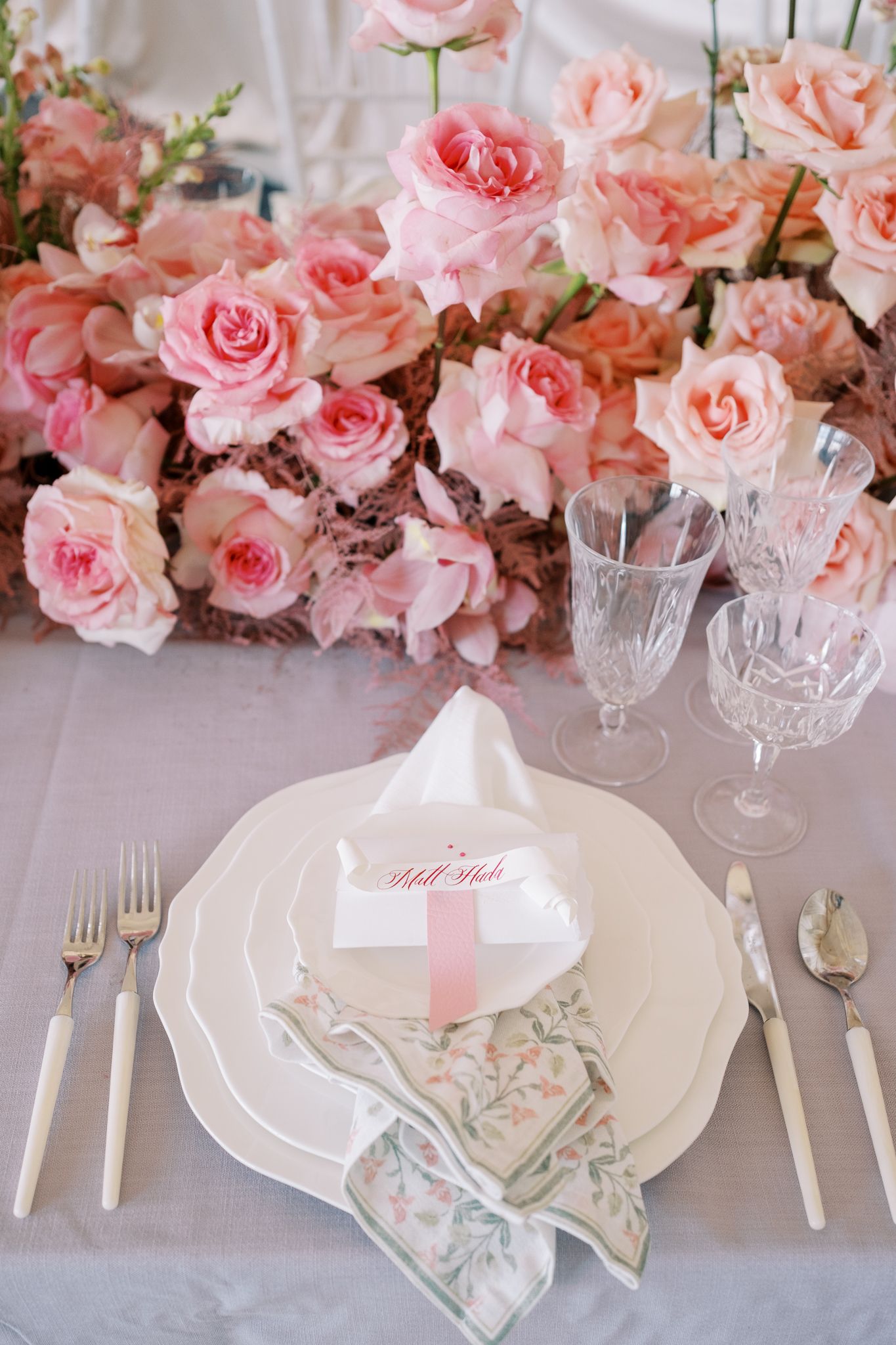 Elegant table setting with pink roses, crystal glasses, and floral patterned napkins on a light gray tablecloth.