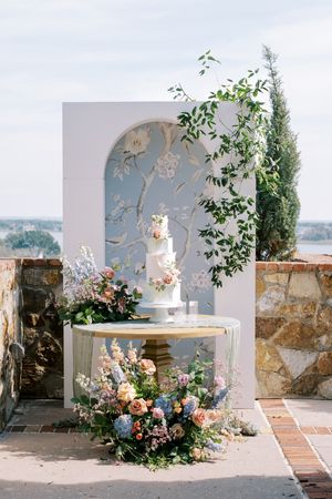 Elegant wedding cake display with floral arrangements against a blue chinoiserie arch backdrop on an outdoor terrace.