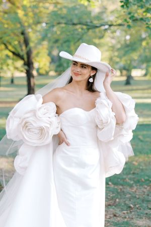 Elegant bridal fashion featuring white gown with dramatic puff sleeves and wide-brimmed hat in garden setting.