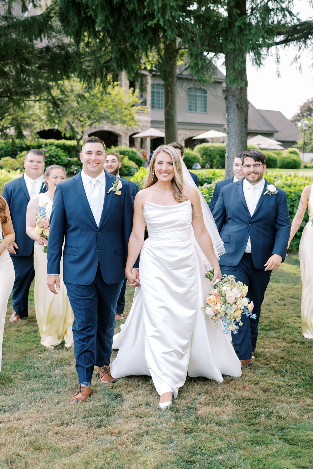 Wedding party in navy suits and white dresses walking and laughing together on grass during outdoor celebration.