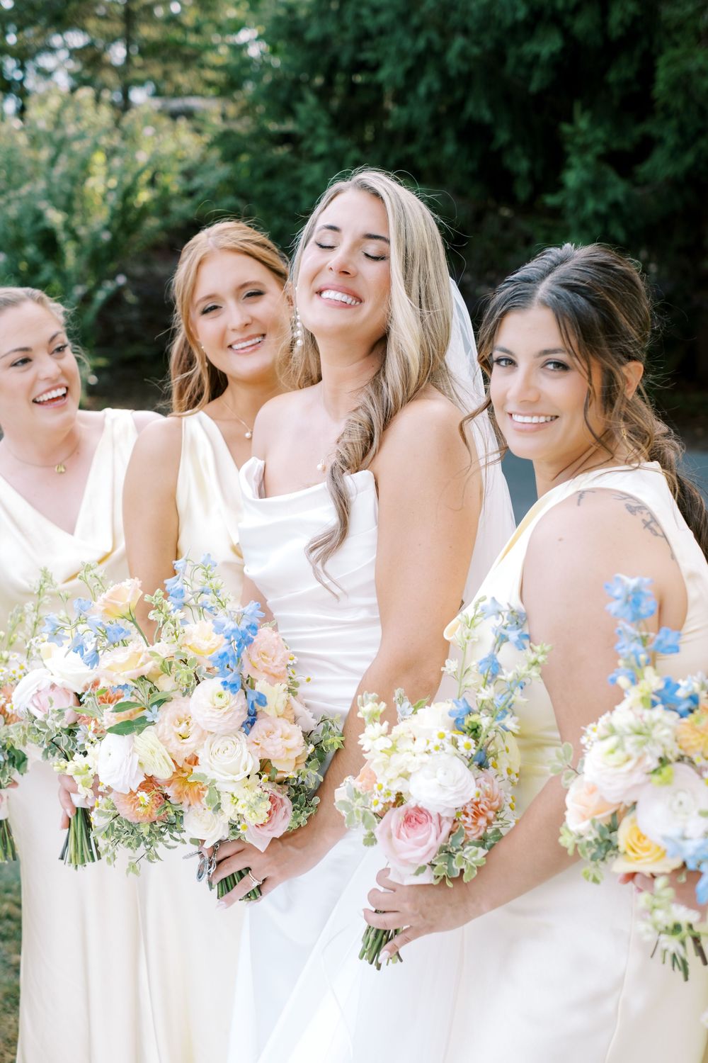 Bridesmaids in white dresses hold colorful bouquets of pink and blue flowers while laughing together outdoors.
