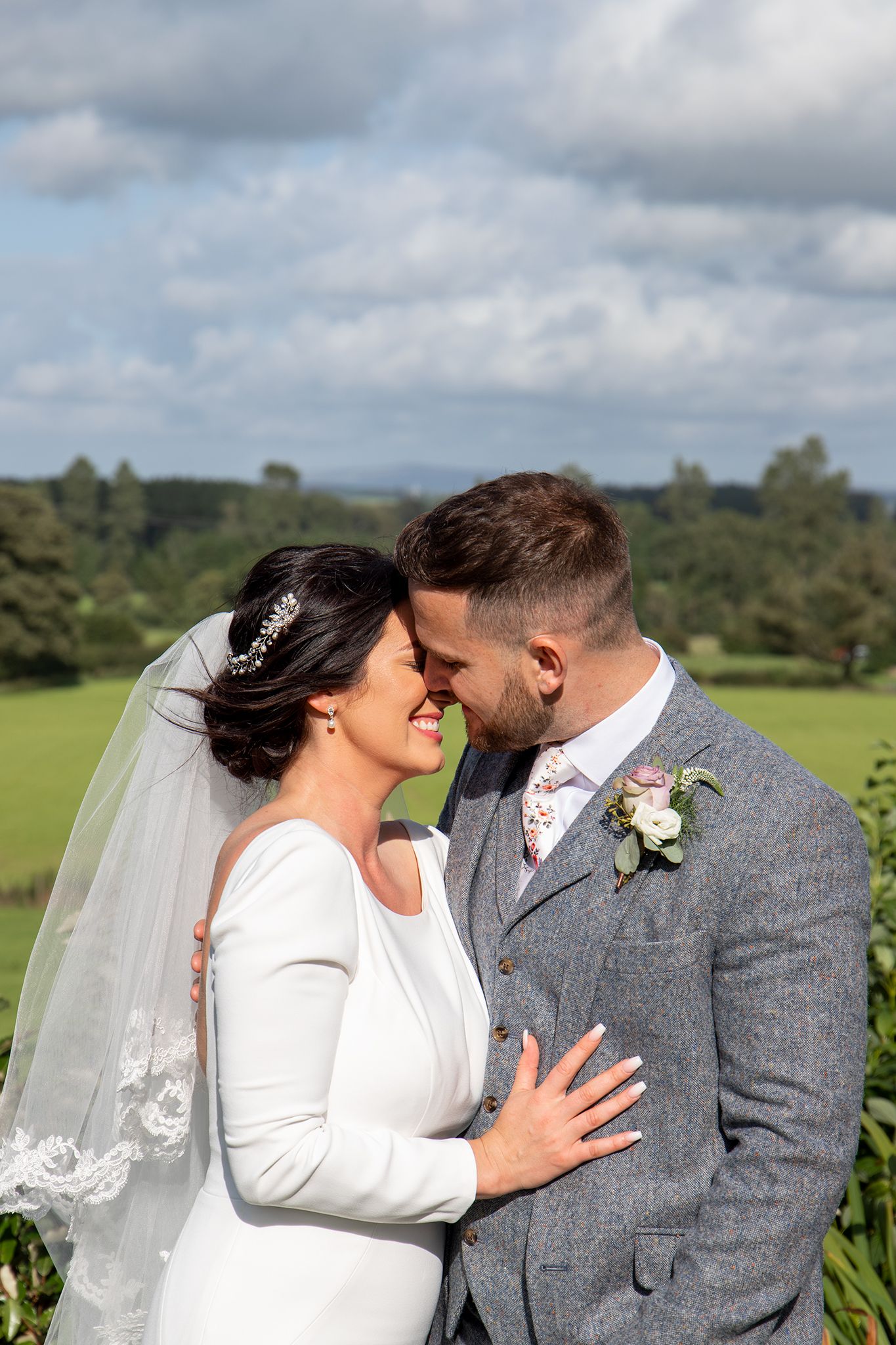 A romantic wedding portrait outdoors with rolling hills and cloudy sky in the background.