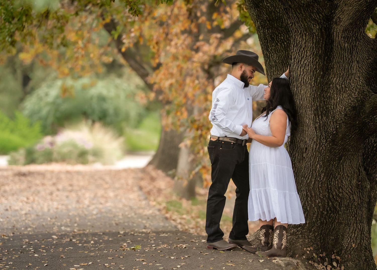 Couple in white dress and cowboy hat share romantic moment leaning against large tree on autumn path.