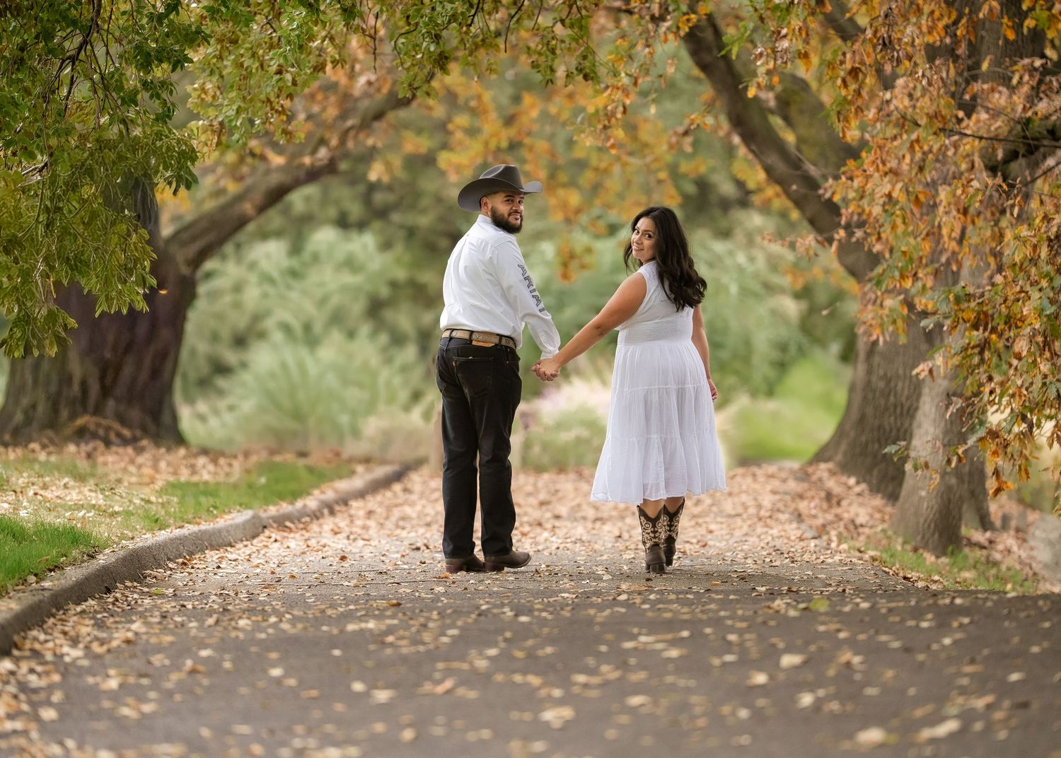 Couple holding hands walking down a tree-lined autumn path, woman in white dress and man in hat.