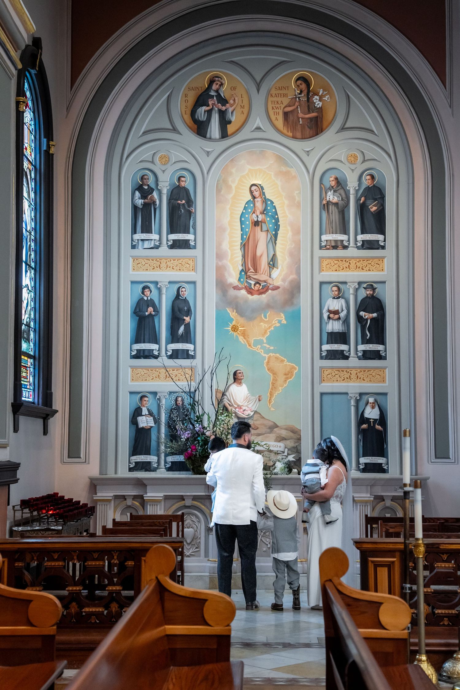 Family standing before Our Lady of Guadalupe mural in ornate Catholic church with saints and stained glass windows.