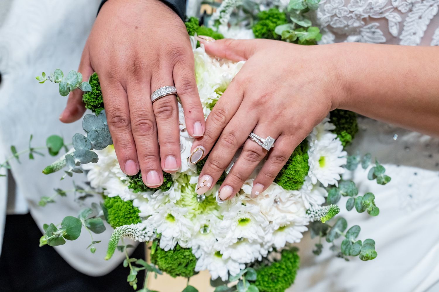Newlywed couple displaying wedding rings on a bridal bouquet of white daisies and greenery.