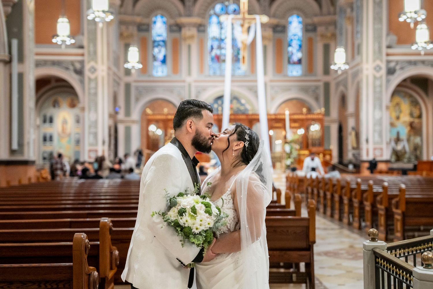 Bride and groom share a kiss in an ornate church aisle, bride holding white bouquet, stunning stained glass windows behind.