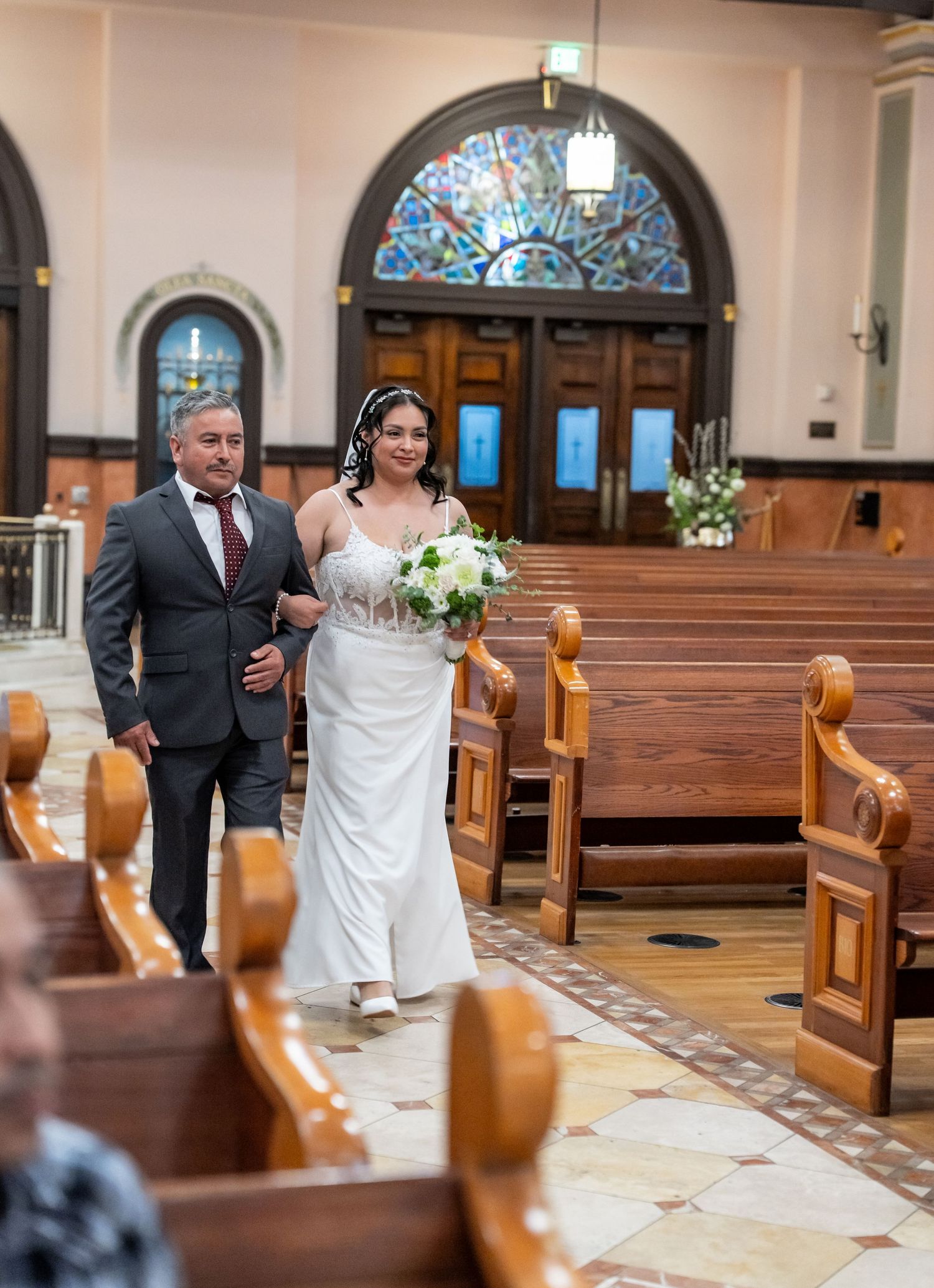 Bride in white gown walks down church aisle with escort past wooden pews and stained glass windows.