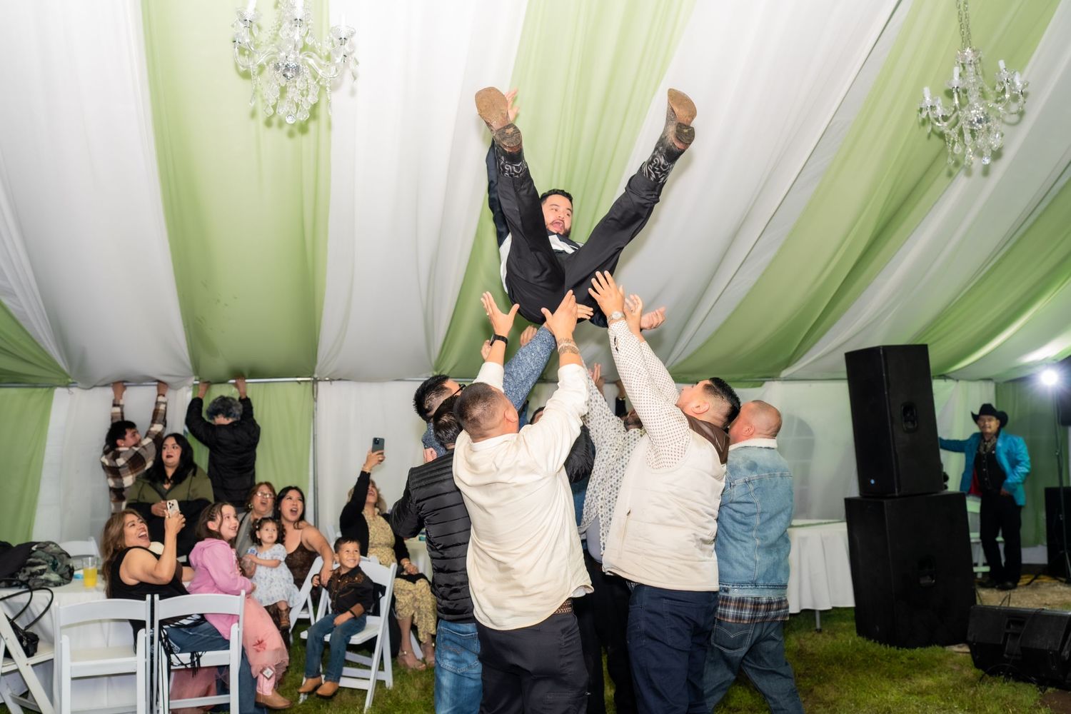 Groomsmen lifting groom in the air at a wedding reception inside a decorated tent with chandeliers.