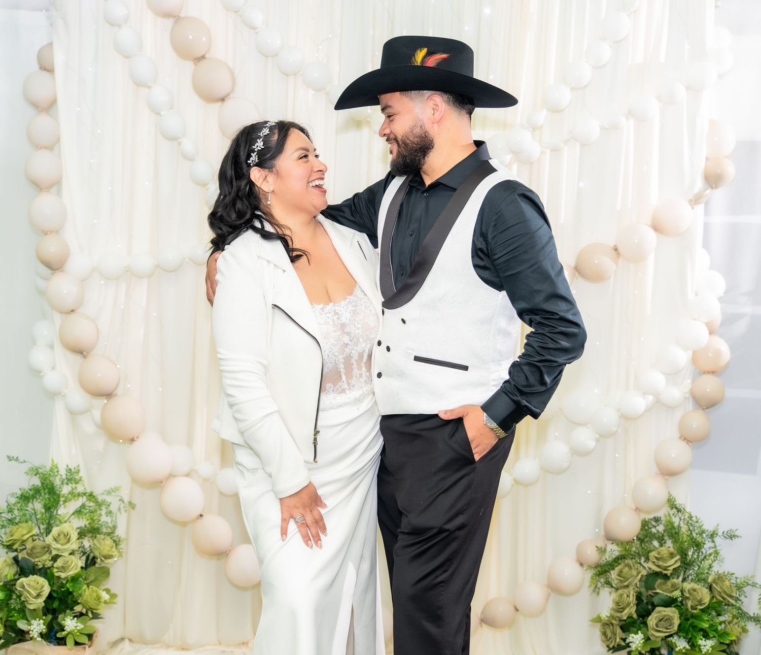 Couple in wedding attire smiling at each other in front of a white balloon arch with floral decorations.