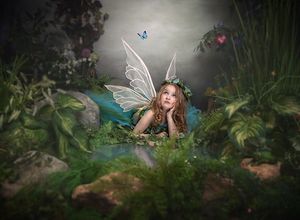 A young girl dressed as a fairy with wings and floral crown rests among lush greenery in an enchanted forest setting.