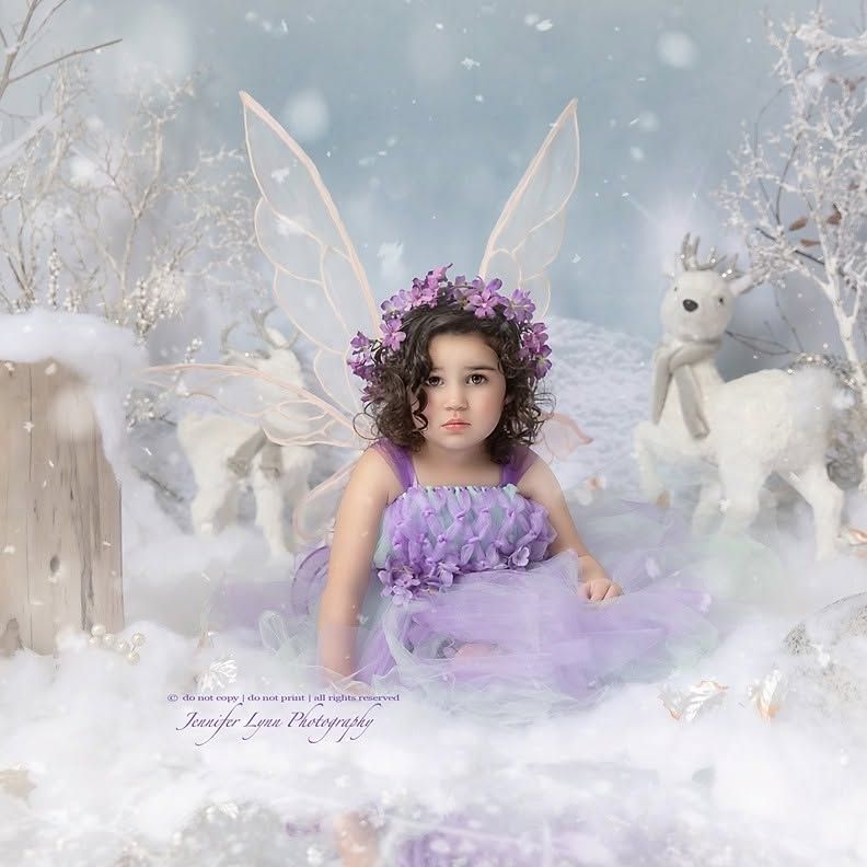 A young girl in a purple fairy dress and floral crown poses in a whimsical winter wonderland with white deer and fairy wings.