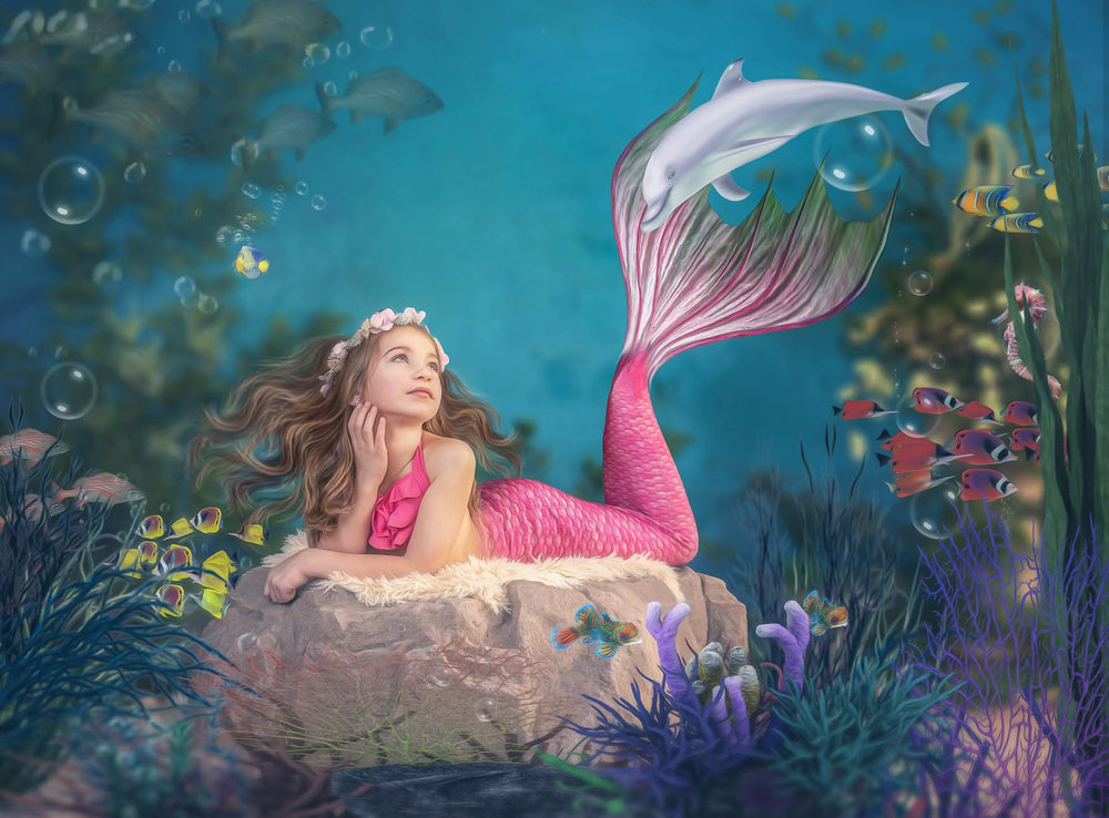 A young girl dressed as a mermaid with a pink tail poses on a rock in an enchanting underwater fantasy scene.