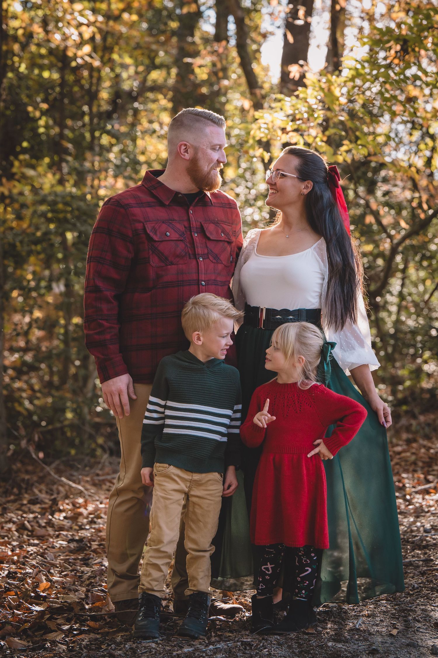 A family in autumn outfits stands together in a forested area with golden fall foliage in the background.
