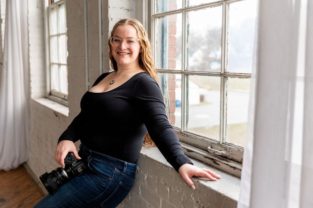 Cheerful woman in black top leans on a windowsill holding a camera in a bright brick-walled studio.