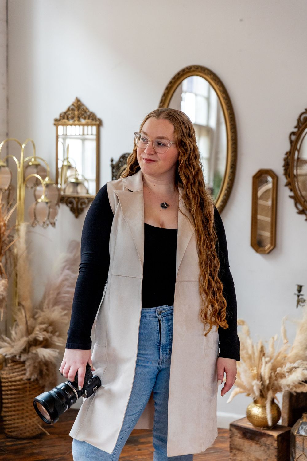 Female photographer with long curly hair holding camera in a boho-styled studio with vintage mirrors.