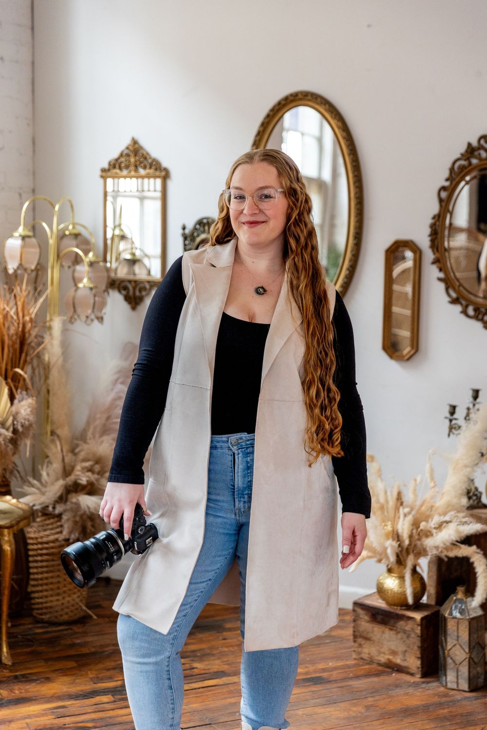 Smiling female photographer in beige vest and jeans posing with camera in a vintage boho studio.