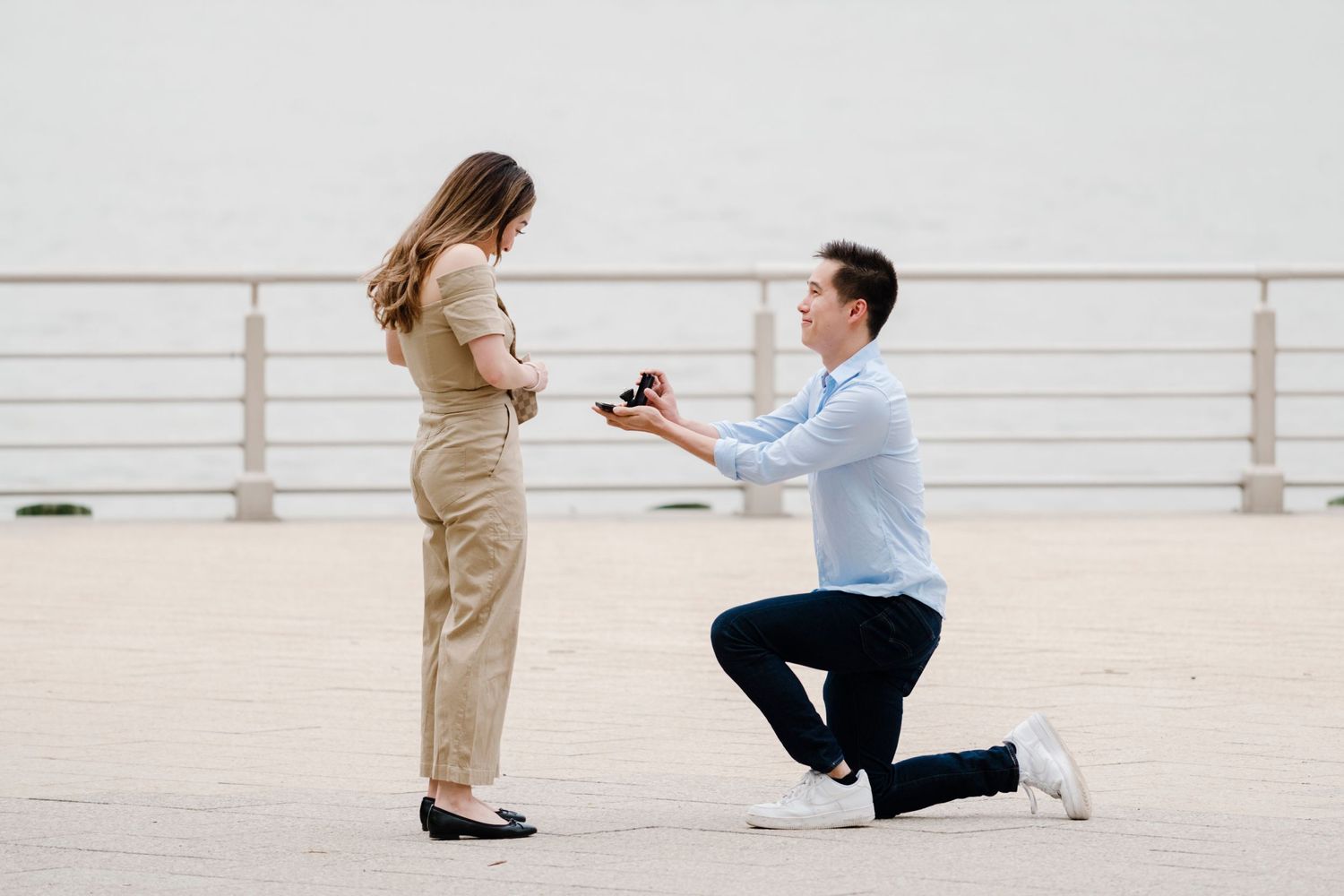 A romantic proposal scene with one person kneeling and holding a ring box on a bright outdoor terrace.
